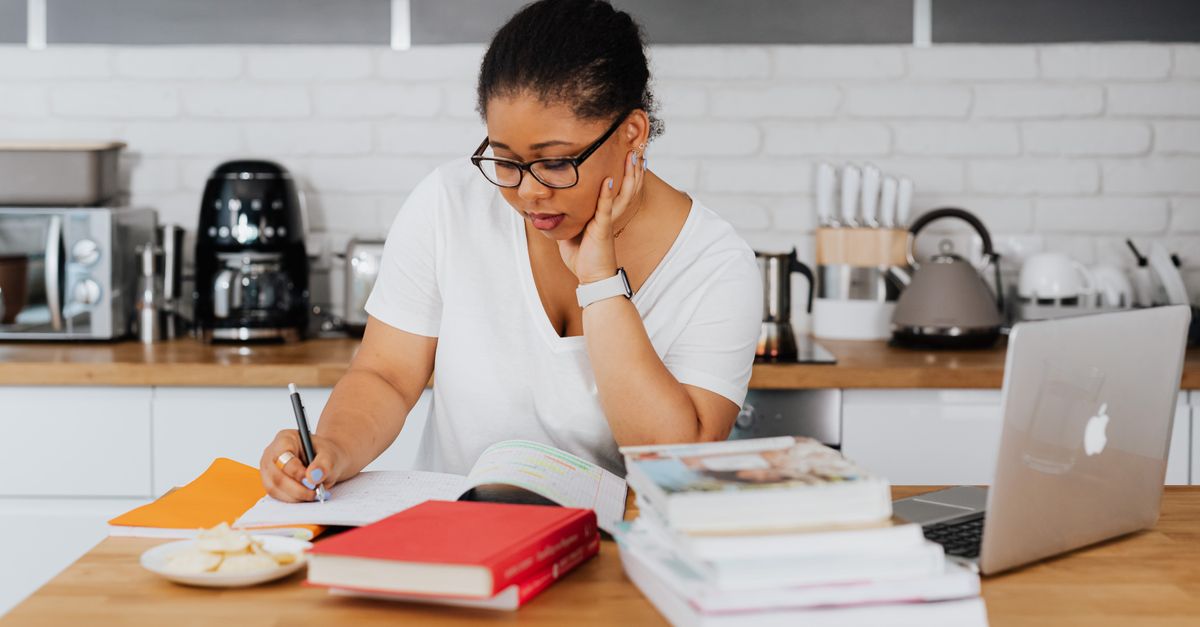 Woman writing at a kitchen table with a laptop and notebooks