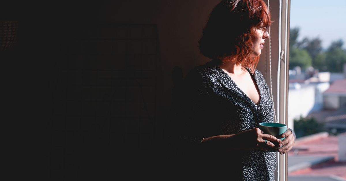 Woman in her forties standing at a window at dusk holding a mug