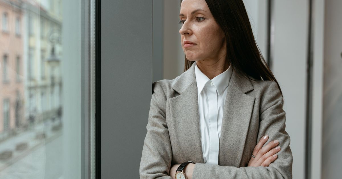 Woman in her forties standing by an office window with her arms folded, looking away